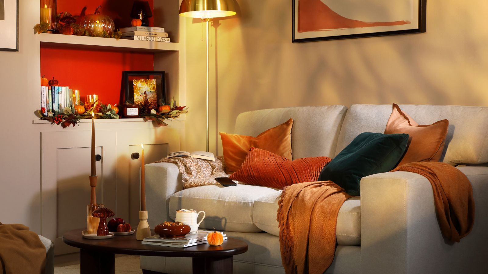 A neutral sofa is layered with warm autumnal cushions and throws, while soft golden lighting and pumpkin-filled shelves create a cosy, seasonal corner.