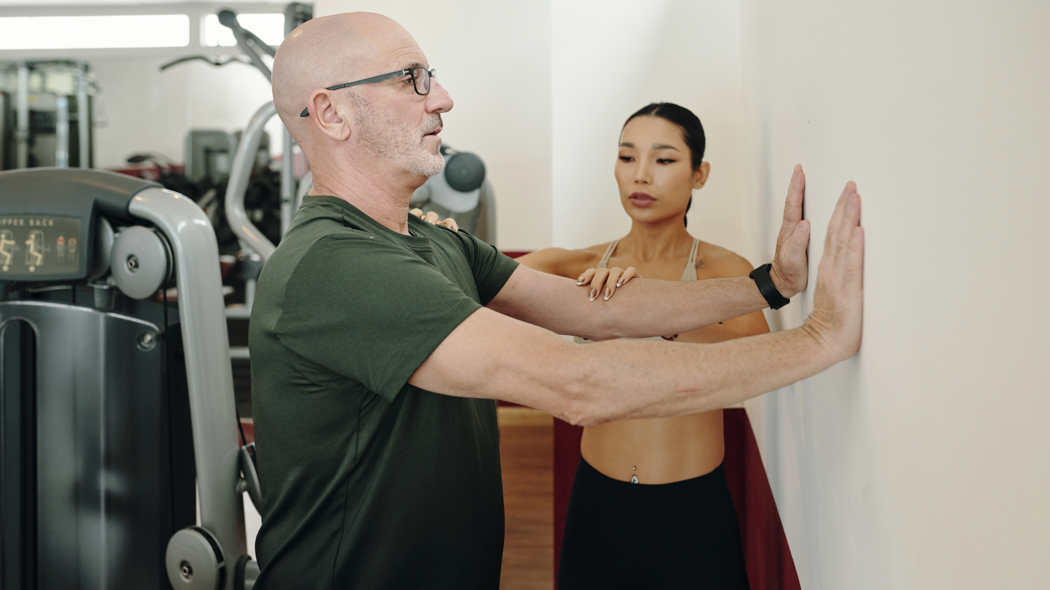 Personal trainer helps adjust the position of an older man who has his hands placed on a wall