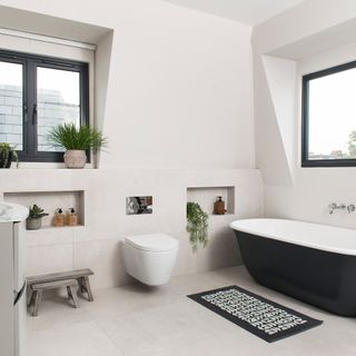A loft bathroom with white wall paint decor, black-framed window, matt black bathtub and various recesses in wall used as storage space