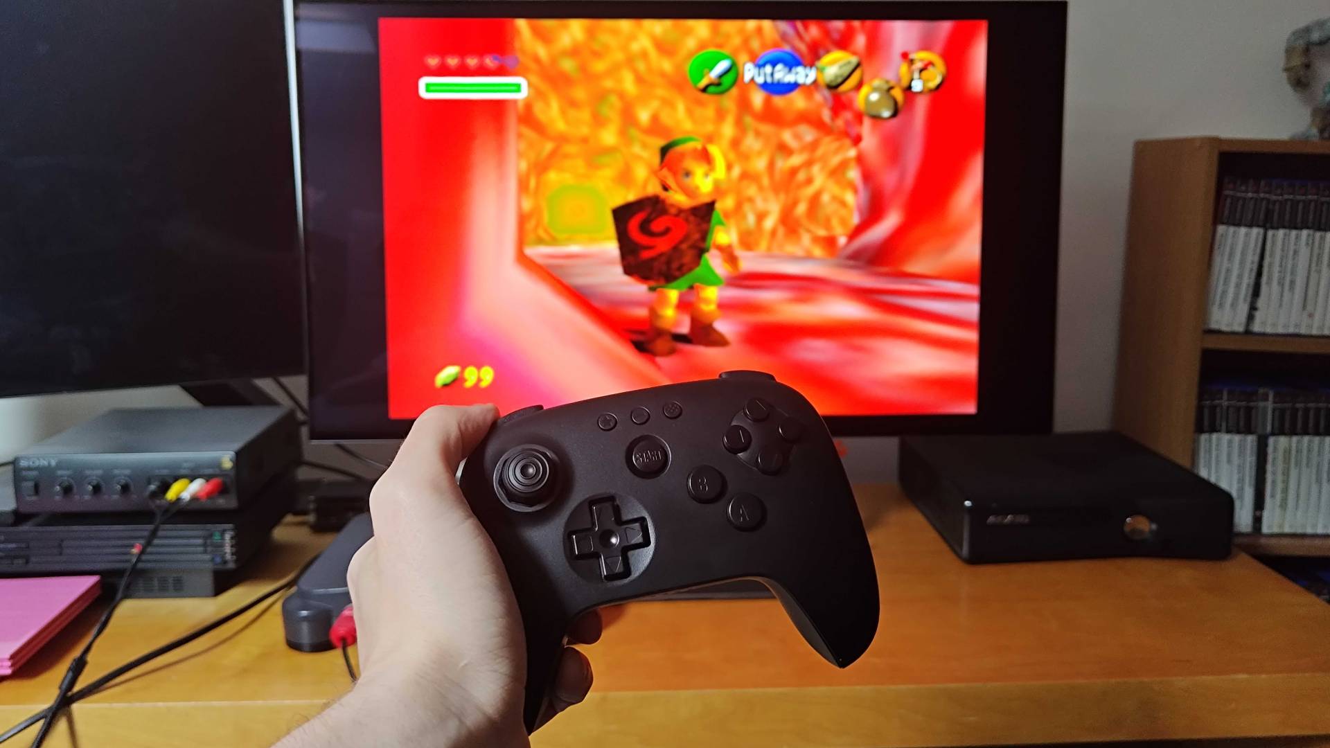 Hand holding 8Bitdo N64 Bluetooth controller with Ocarina of Time gameplay in backdrop on monitor.