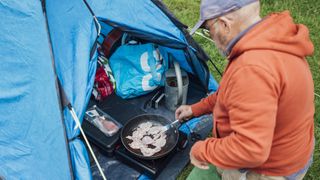 An over the shoulder view of a senior man kneeling down by his tent to cook bacon in a portable camping stove, he is preparing breakfast at his campsite at Hollows Farm, The Lake District in Cumbria, England