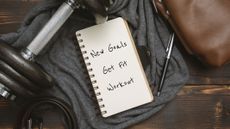 A picture of a notebook lying next to a dumbbell, pen and gym towel, with the front page having "New goals: Get fit, workout" written on it 