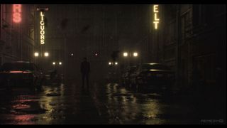 Eine stimmungsvolle Stadtlandschaft. Wasser türmt sich auf dem Boden, während Neonschilder die dunklen Straßen erhellen. Alan Wake steht in der Mitte des Bildes.