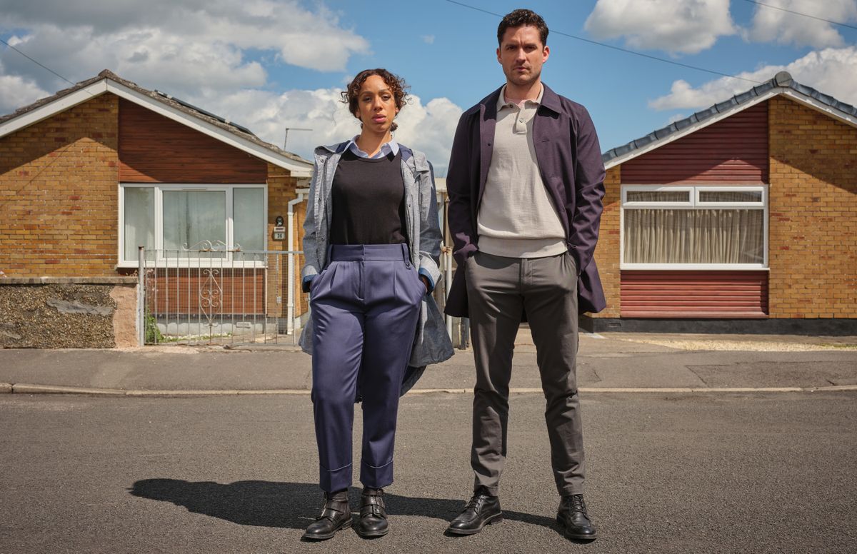 Pearl Mackie as DS Jen Rafferty and Ben Aldridge as DI Matthew Venn in The Long Call, both standing with their hands in their pockets on the street in front of a row of bungalows