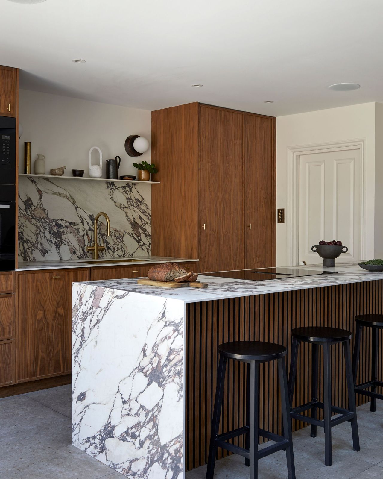 A modern kitchen with a marble-topped kitchen island with reeded wood detailing on the side with black bar stools that match the wood used for the cabinetry on the main kitchen wall behind