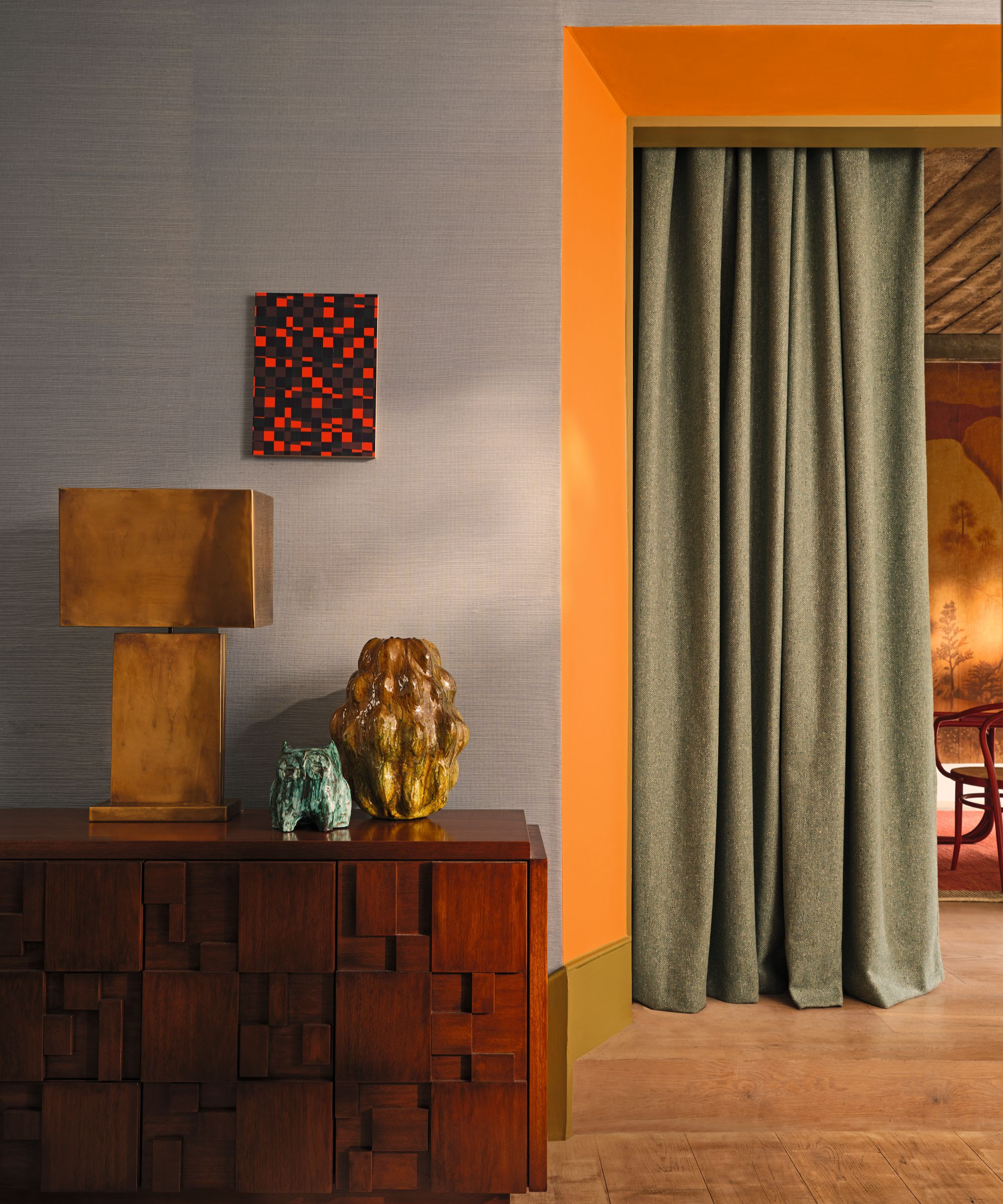 a room with a wood sideboard, a gold lamp, a vase, a small painting on the wall, grasscloth wallpaper, an orange hallway, and a curtain separating the hallway from another room