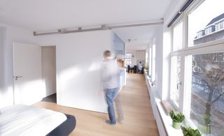 A room with white walls, ceilings and wooden floor. On the left is a double bed with white sheets and blue base and an open white door. On the right is a corridor that leads to other room with clear glass window panels on the right of the corridor