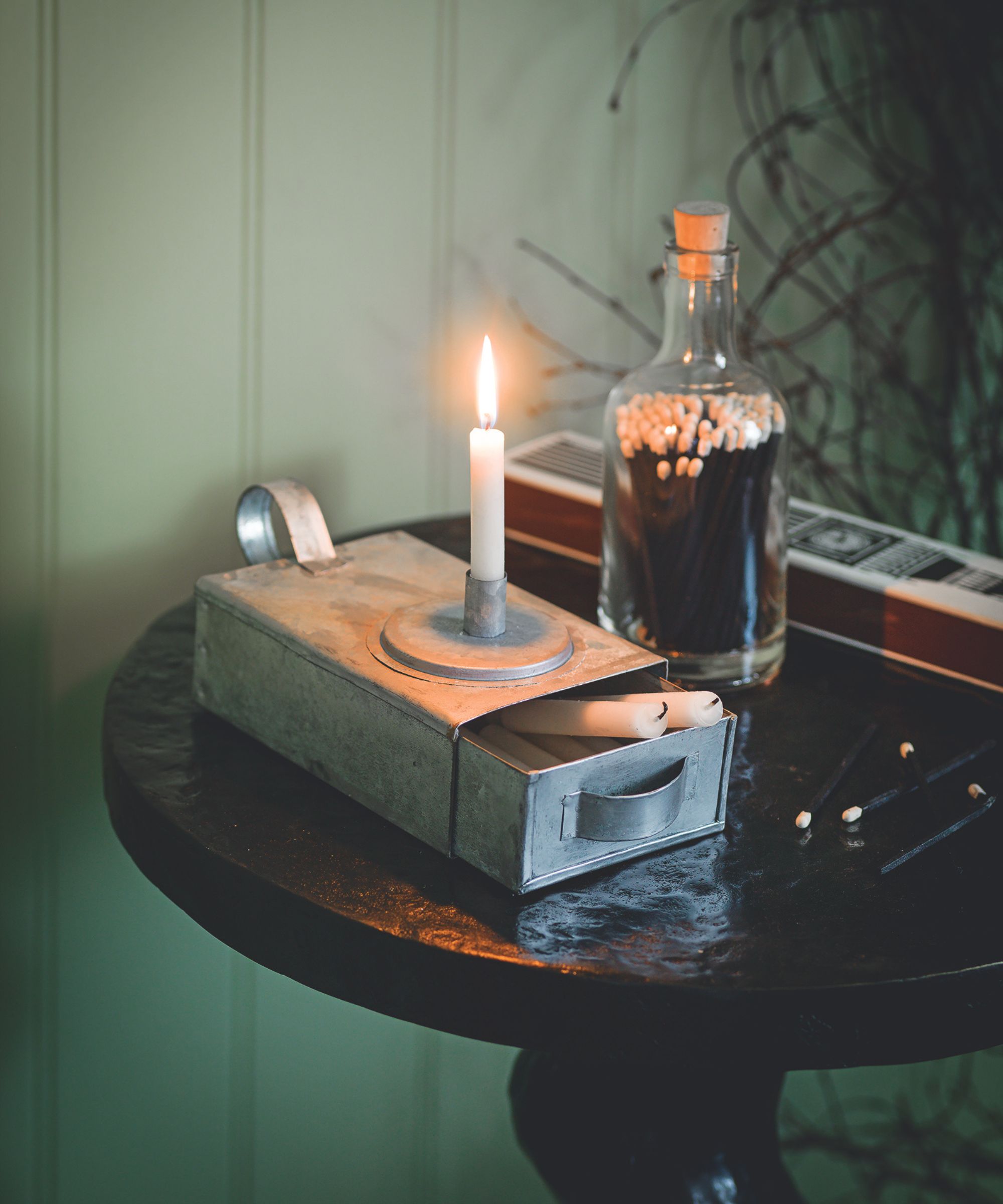 White taper candle in metal holder with matches in a glass bottle on a wooden circular table. The wall behind is sage green and panelled.