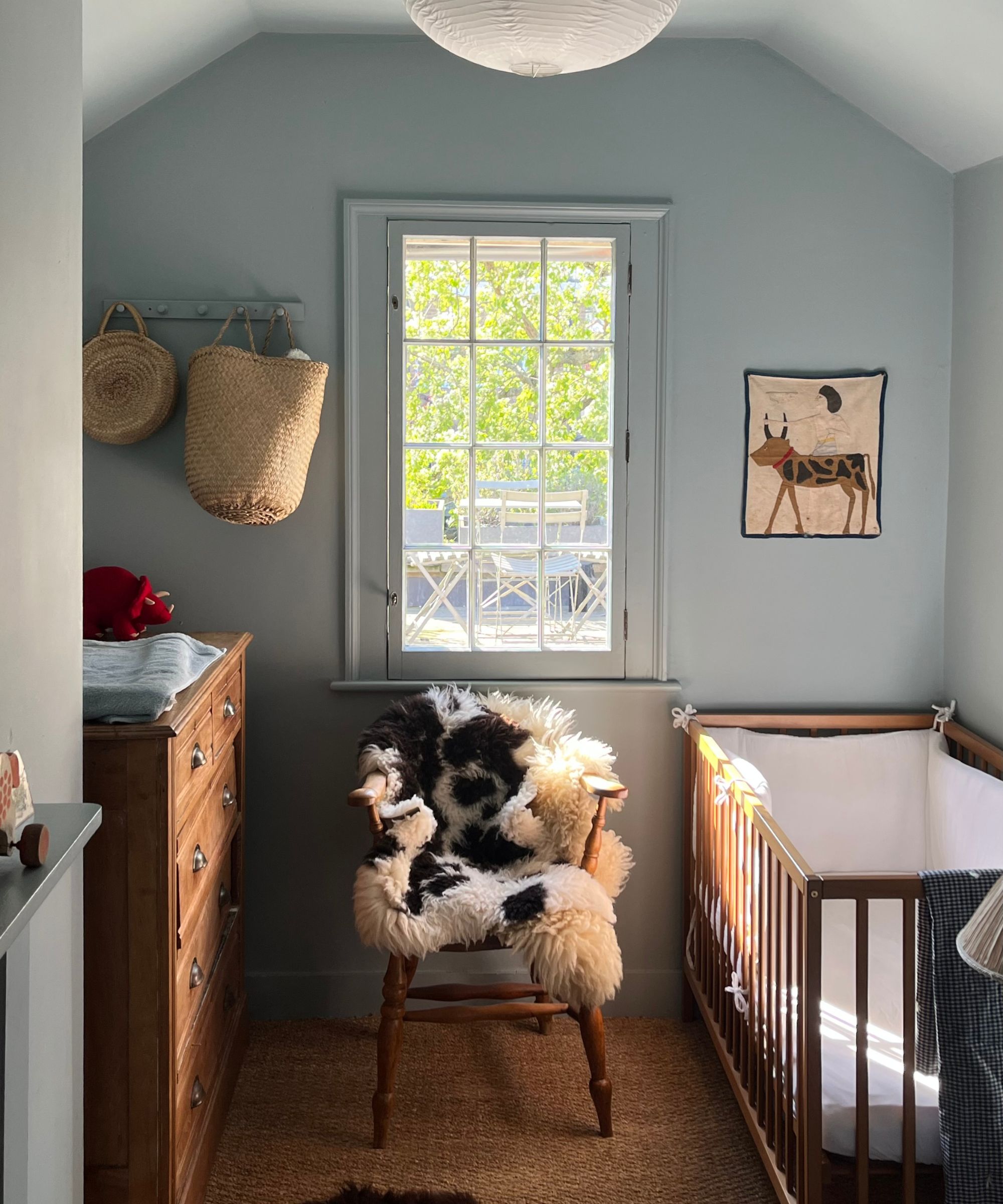 Nursery with pale blue painted walls, rattan carpet, wooden dresser, vintage wooden chair with sheepskin throw, a wooden cot and baskets hanging on coat hooks