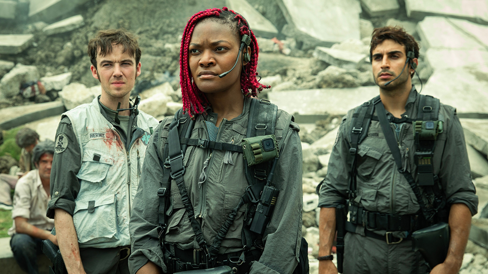 Still from the TV show Alien: Earth. Three people are standing together in front of a large pile of rubble. They are each wearing a gray jumpsuit, utility belt, backpack, and a headset with a mic.