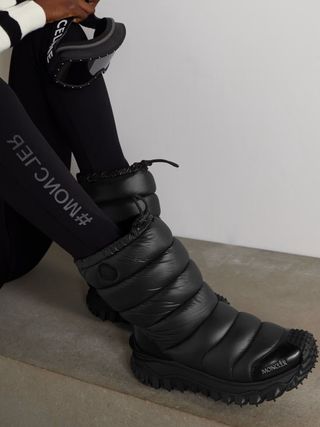 A woman dressed in ski gear wears tight leggings in black that read "#MONCLER" and a pair of chubby black books with spiky ends.