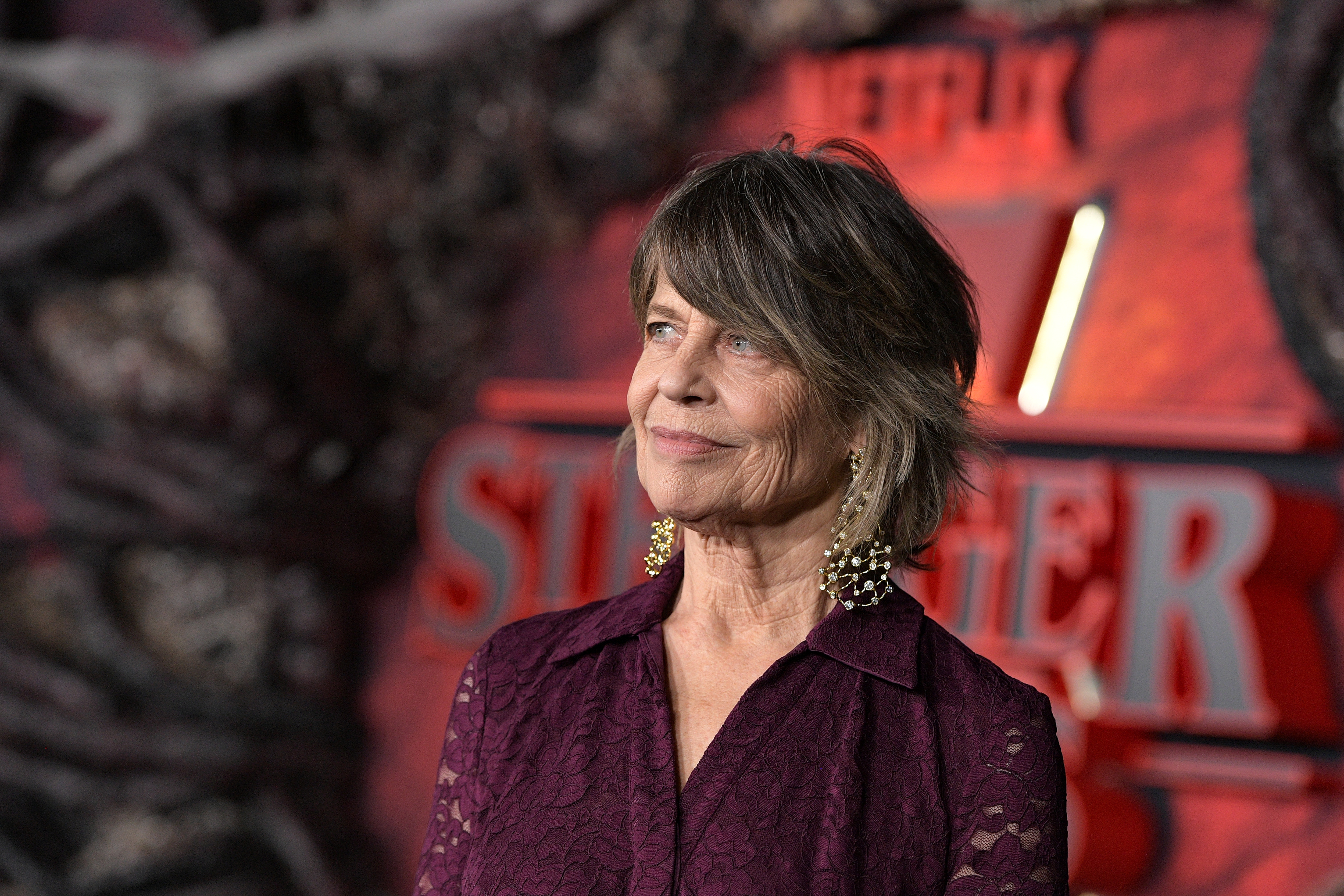 actress linda hamilton smiles and poses on the red carpet for the stranger things season 5 premiere waring a purple lace dress and large earrings
