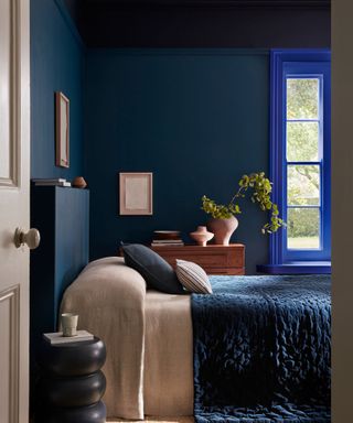 Blue bedroom with bright blue window frame, bed with blue quilt and wooden drawers with vase and foliage