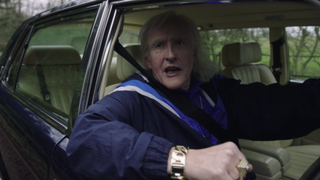 Jimmy Savile (Steve Coogan) dressed in a trademark blue tracksuit and leaning out the window of his Bentley car.