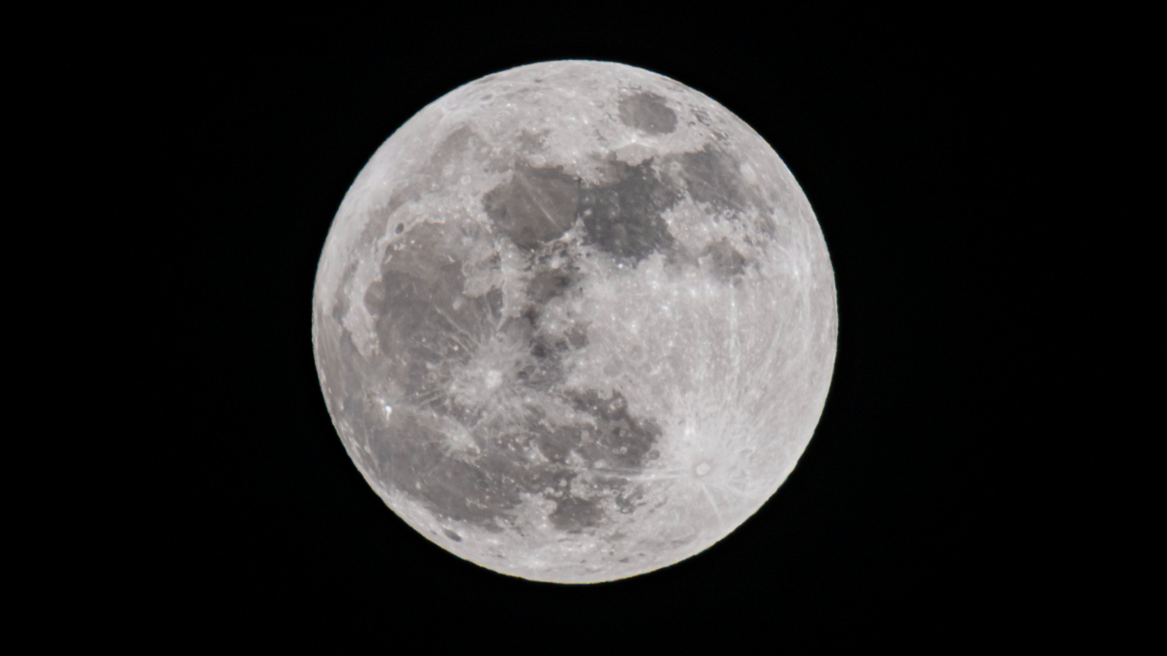 A full moon is pictured shining against a black night sky as the dark forms of lunar maria darken the lunar surface where ancient lava flows have cooled and solidified.