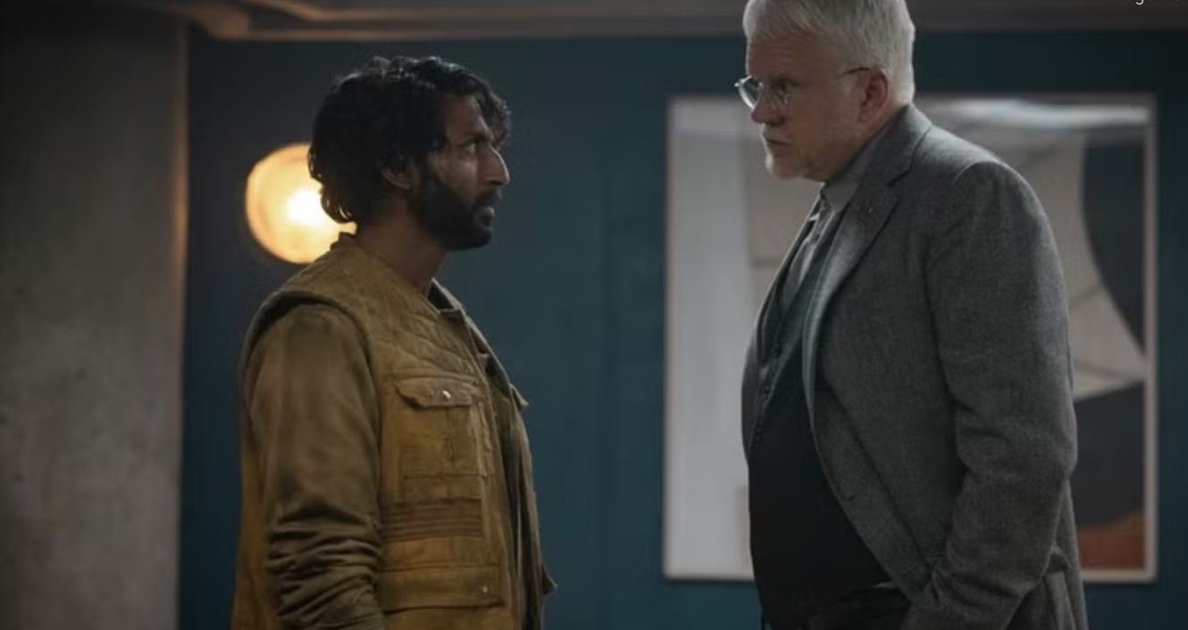 A bearded man in a yellow vest stands opposite a tall man with white hair