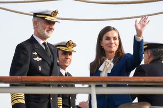 Queen Letizia wearing a blue suit and white tie-neck blouse waving from a ship standing next to King Felipe in a naval uniform