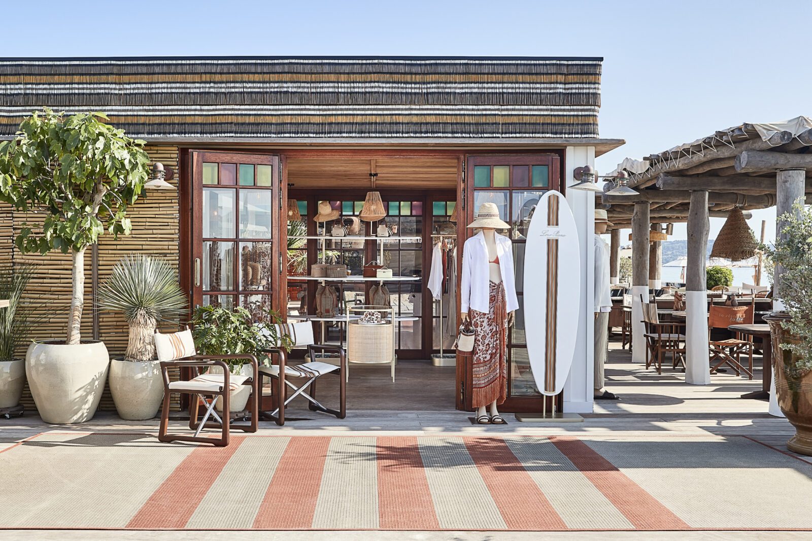 A resort-themed, design-forward fashion beach club features minimalist, stripy decor in hues of beige, coral, and white and a "cabana"-style, flat-roofed storefront.