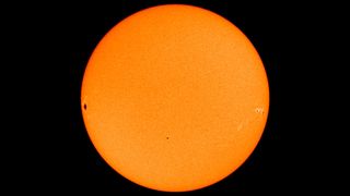 The planet Mercury transiting in front of the sun, as captured by a NASA satellite. Mercury is near the center of the image, while dark sunspots appear near the edges.