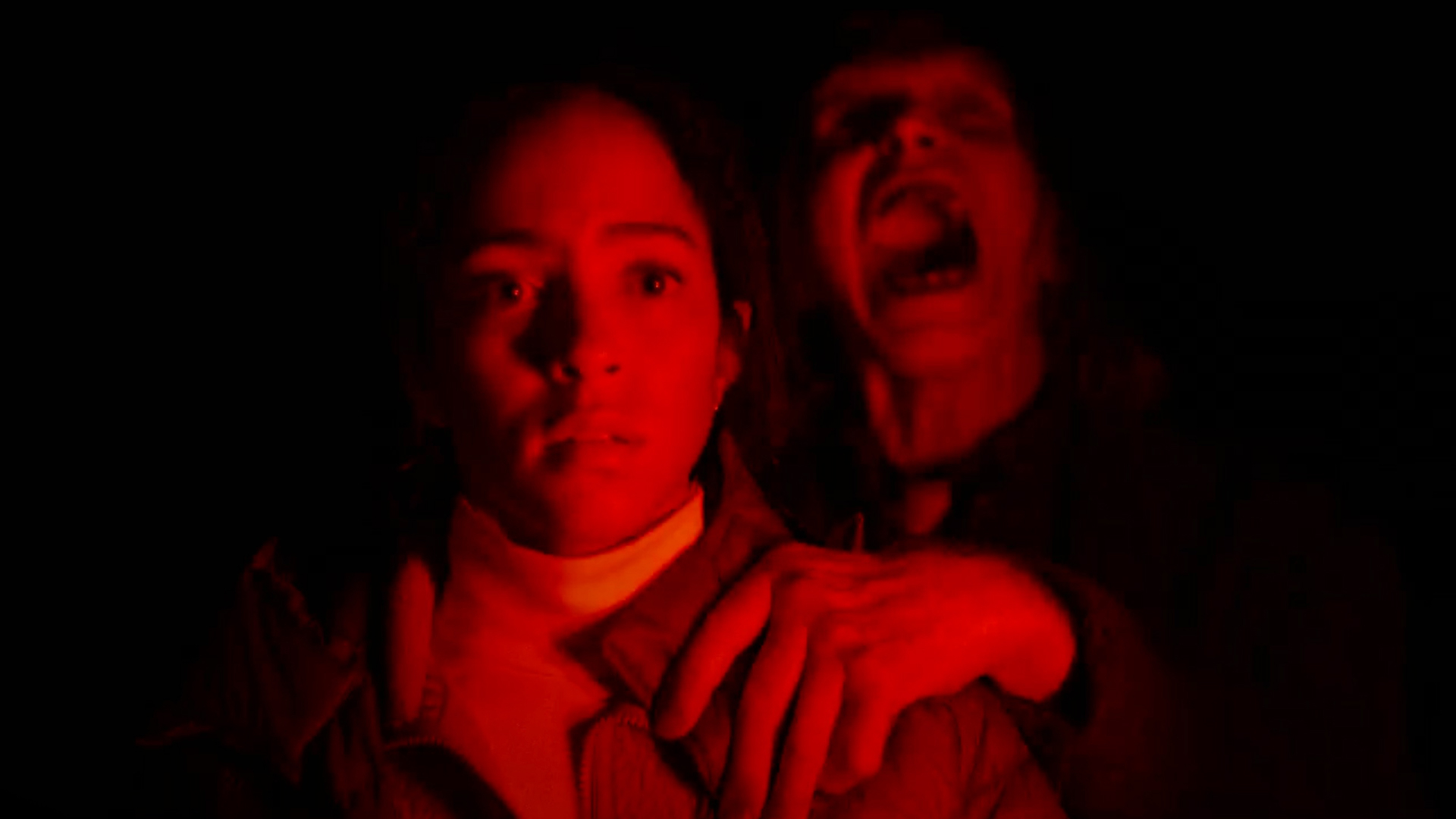 A woman is sitting in a car illuminated by red light. A screaming figure grabs her shoulder behind her.