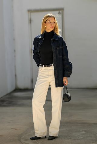 A black turtleneck outfit is shown in a photo of a woman standing on the sidewalk wearing a denim button-down over a black turtleneck with a black belt, white jeans, black ankle boots, and a black handbag