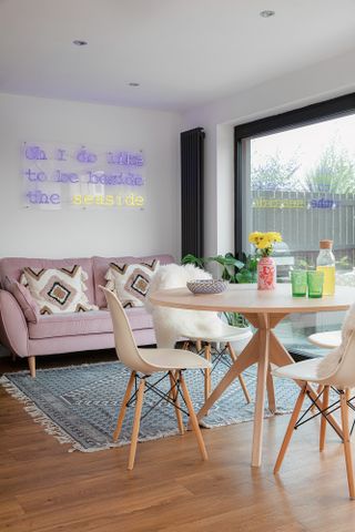 Area of open-plan kitchen-diner with round oak dining table and chairs, blue pattern rug, pink velvet sofa, neon light reading 'Oh I do like to be beside the seaside' and sliding doors opened up to garden