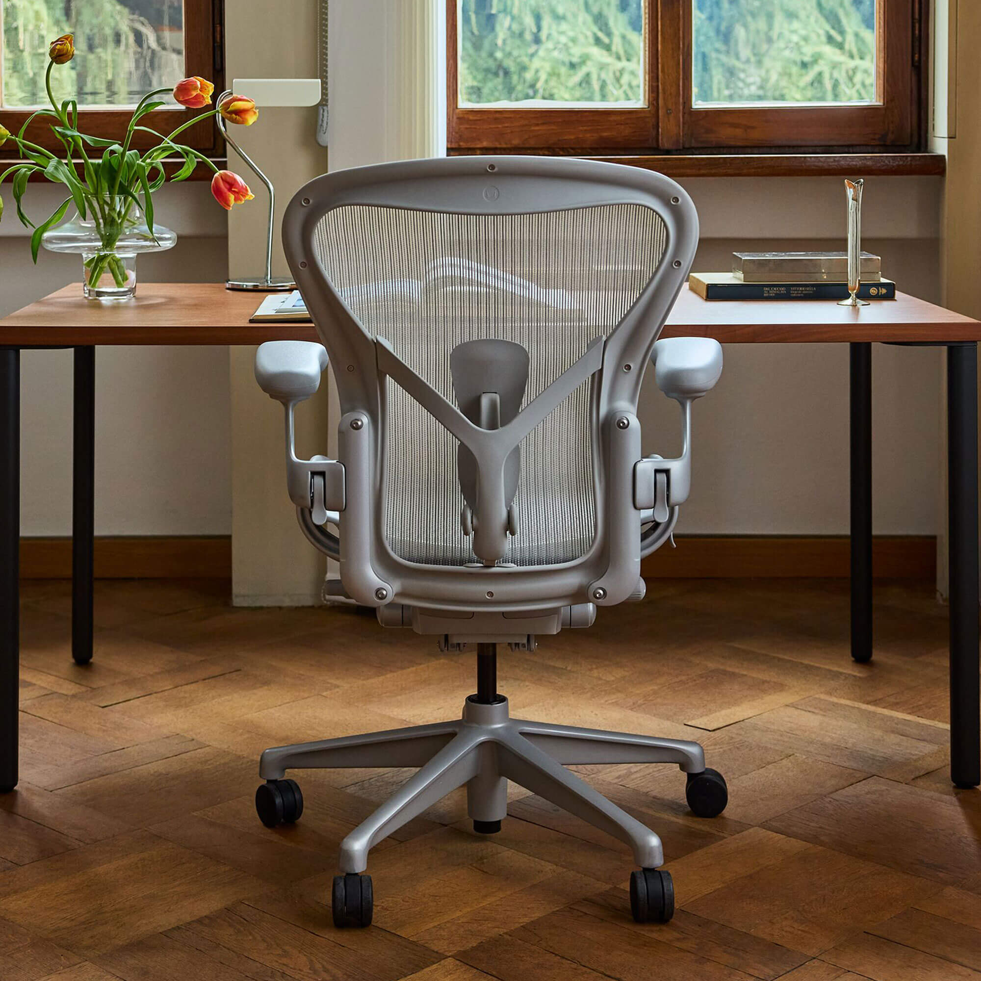 Light grey office chair on wooden floor at wooden desk with vase of tulips in front of windows looking out onto tree