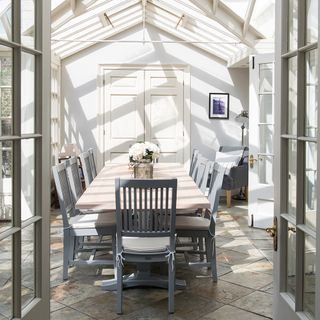 A long dining table with grey chairs in a conservatory with a pitched roof