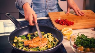 man cooking salmon in a frying pan with courgettes and asparagus 