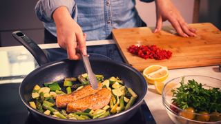 woman cooking salmon in a frying pan with courgettes and asparagus