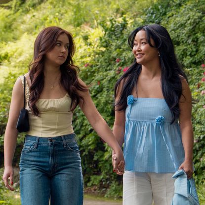 anna cathcart as kitty and lana condor as lara jean holding hands as they walk down a lush path full of greenery and flowers in xo kitty season 3