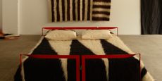 A design showroom hosts textile pieces including an abstract black and white, stripy tapestry, hanging on the wall, and a bed with red iron detailing covered in a similar fabric creation, but with a zig-zag pattern.