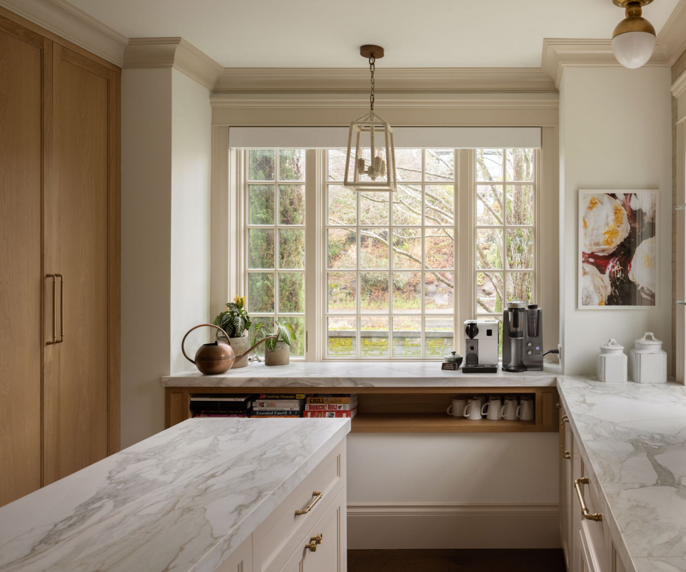 A neutral kitchen with white perimeter cabinets, wooden full-length cabinetry, marble countertops, and a custom cup niche below the countertops bordering the large window