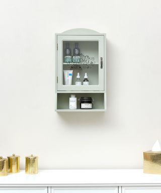 A mint green medicine cabinet with skincare and haircare bottles in it, mounted on a gray wall with a white shelf underneath with three gold containers and a gold tissue box