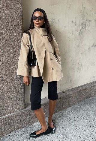 a photo showing the fall jacket trend, funnel neck trench coats worn by a woman standing against a tan wall wearing a tan cropped funnel neck trench coat with black capri pants, black ballet flats, and a black frame handbag