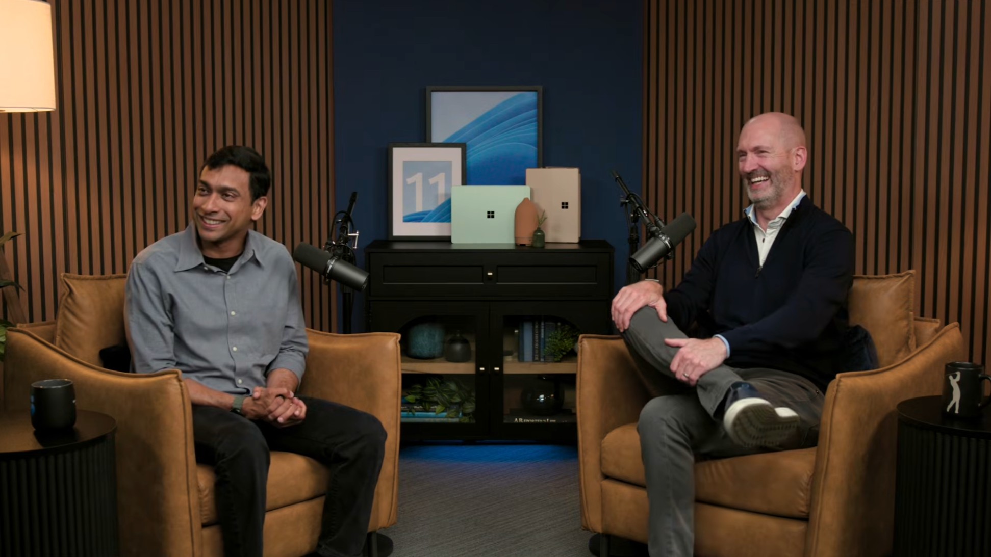 Pavan Davuluri and Brett Ostrum sitting in two arm chairs in front of a display of Surface PCs.