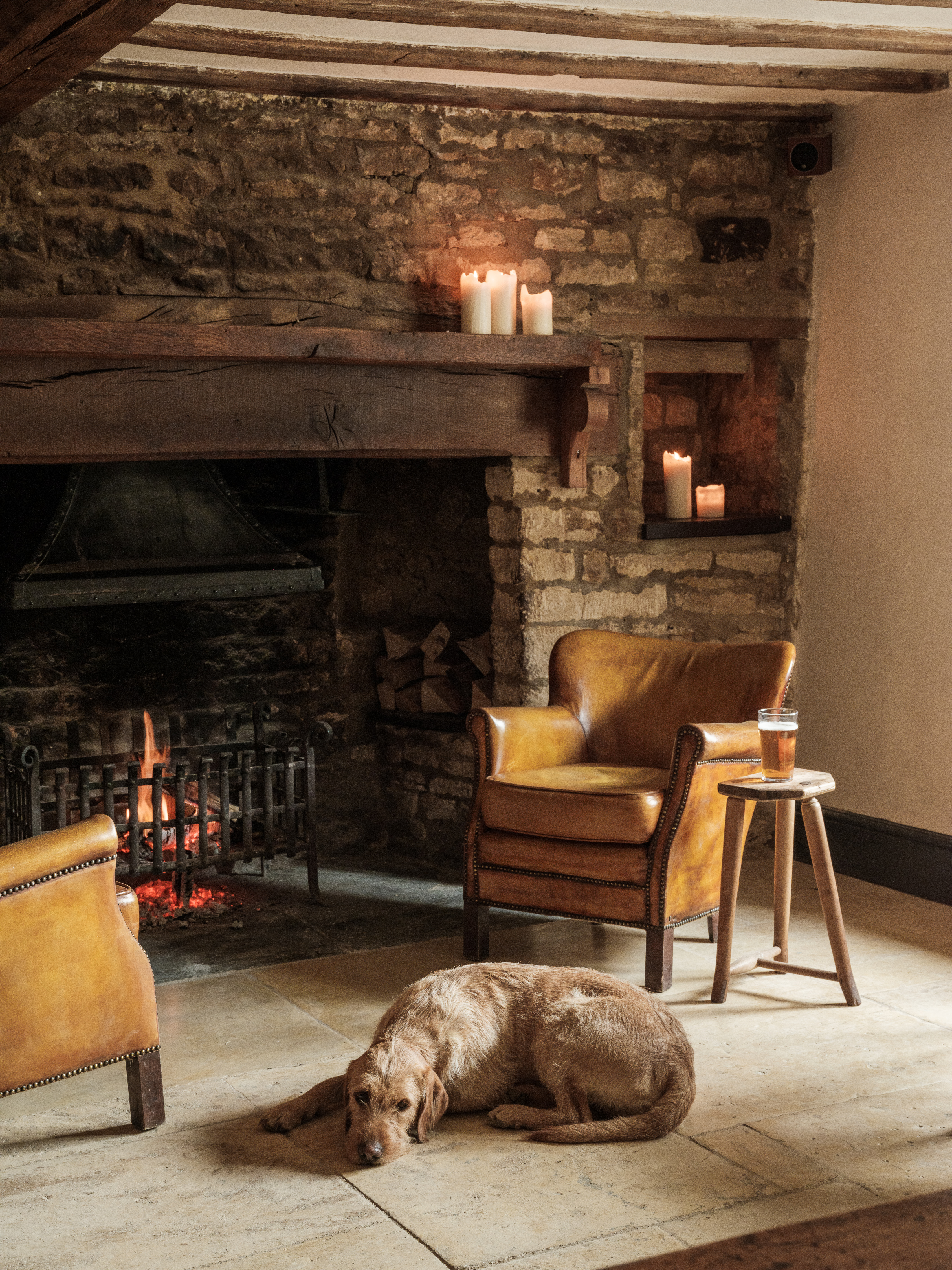 A dog lying down on the ground by a fire at the Bull in Charlbury, surrounded by two armchairs and a pint