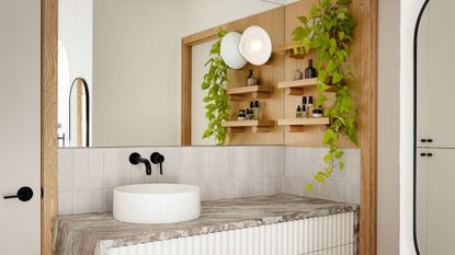 bathroom vanity with full wall length mirror, round sink with black taps, white fluted vanity with grey marble worktop, wood shelves with plants on wall
