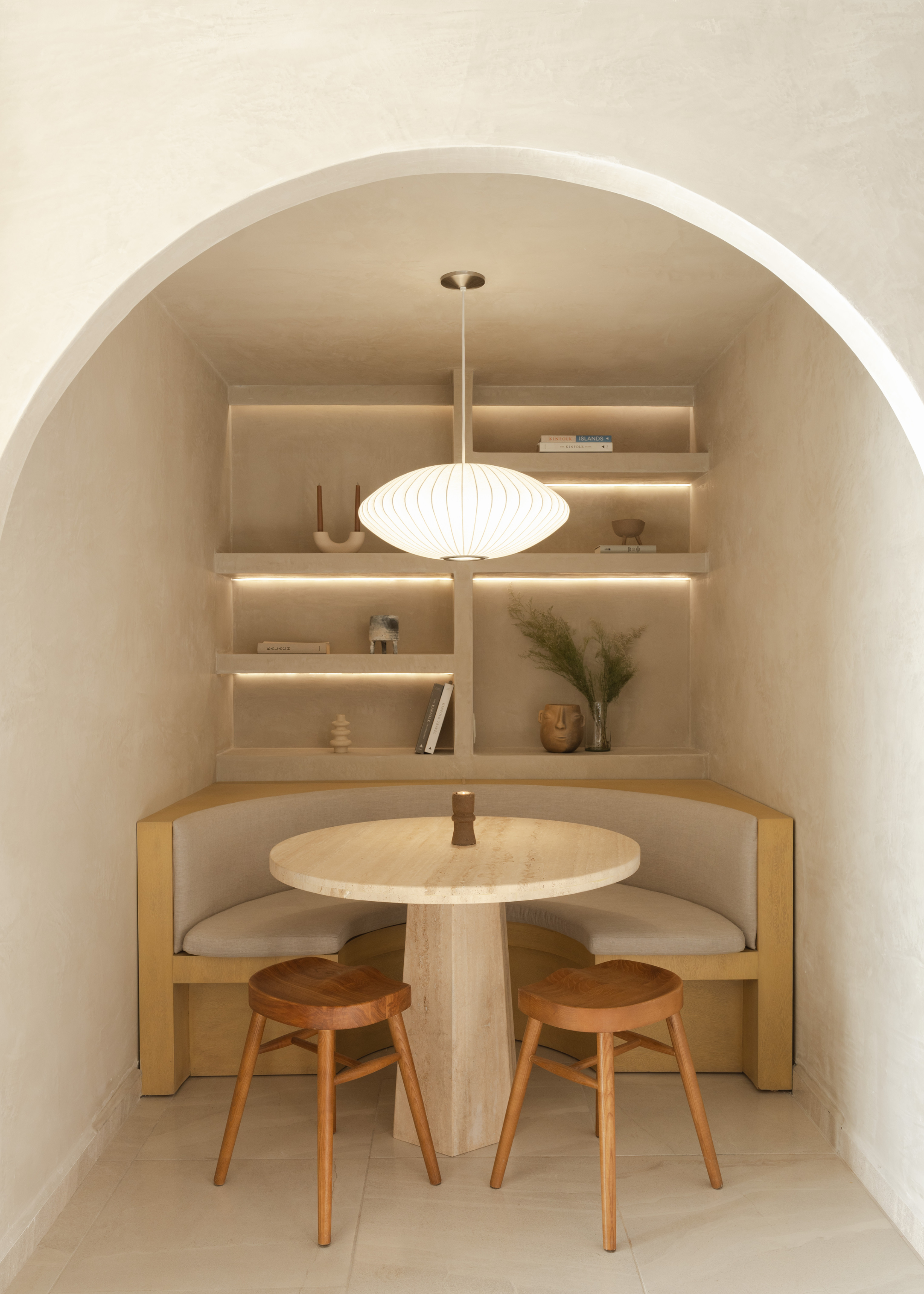 A textured dining nook with off white walls, a shelving unit with candles and books, and a curved bench with a circular stone table, wood stools, and a floating pendant paper lamp