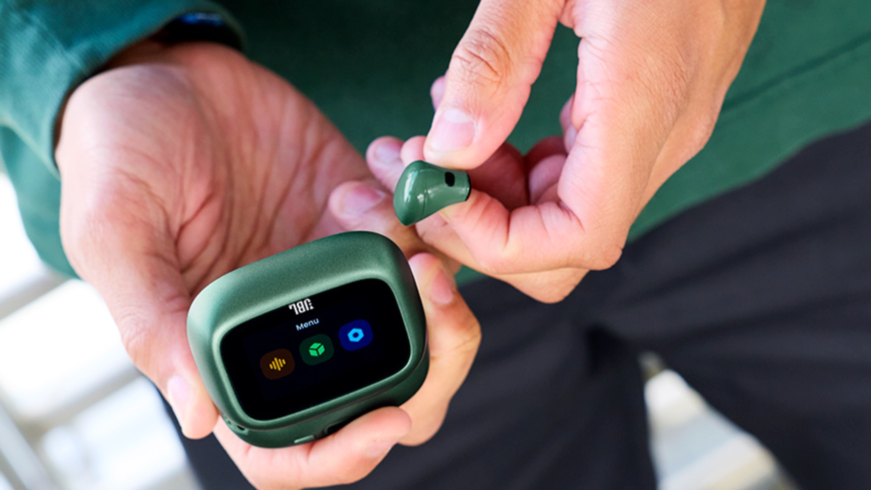 A green JBL Live 4 wireless earbud and charging case held by someone wearing a green shirt.