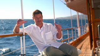 Nick Lachey in a white button-down shirt and grey slacks leaning against the rails of a boat in Perfect Match