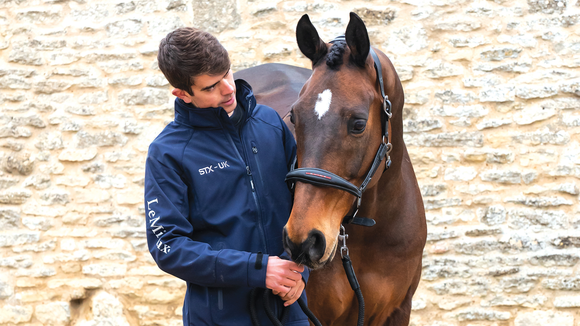 Elite eventing groom Adam Short wears a blue bomber jacket as he stands alongside one of Tom McEwen's bay event horses