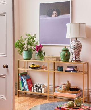 A gold console table with a lamp, plants, and book on it, with a purple wall art print above it