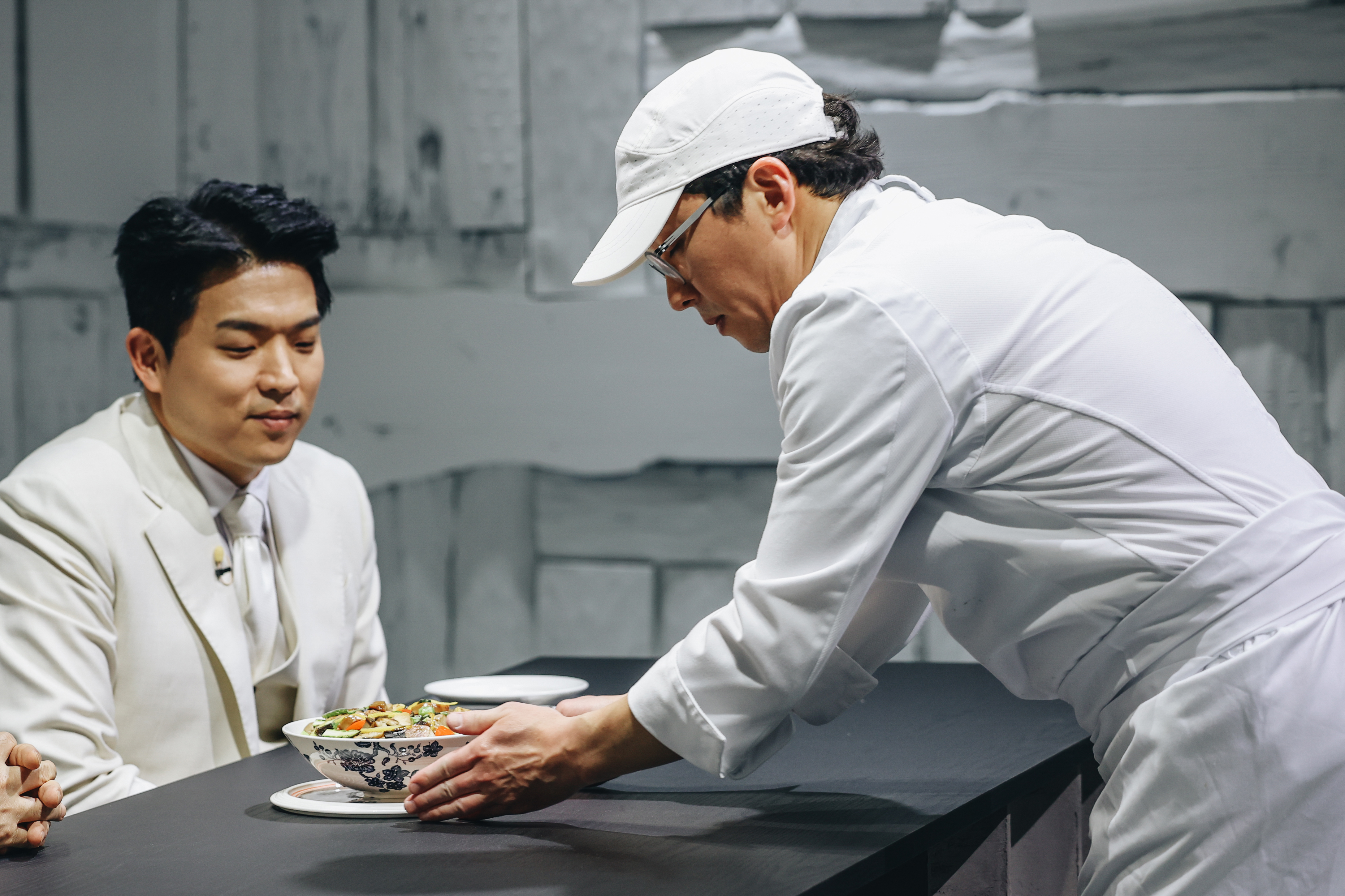 a chef in a white suit and white baseball hat presents a dish to the judges table in a still from culinary class wars season 2