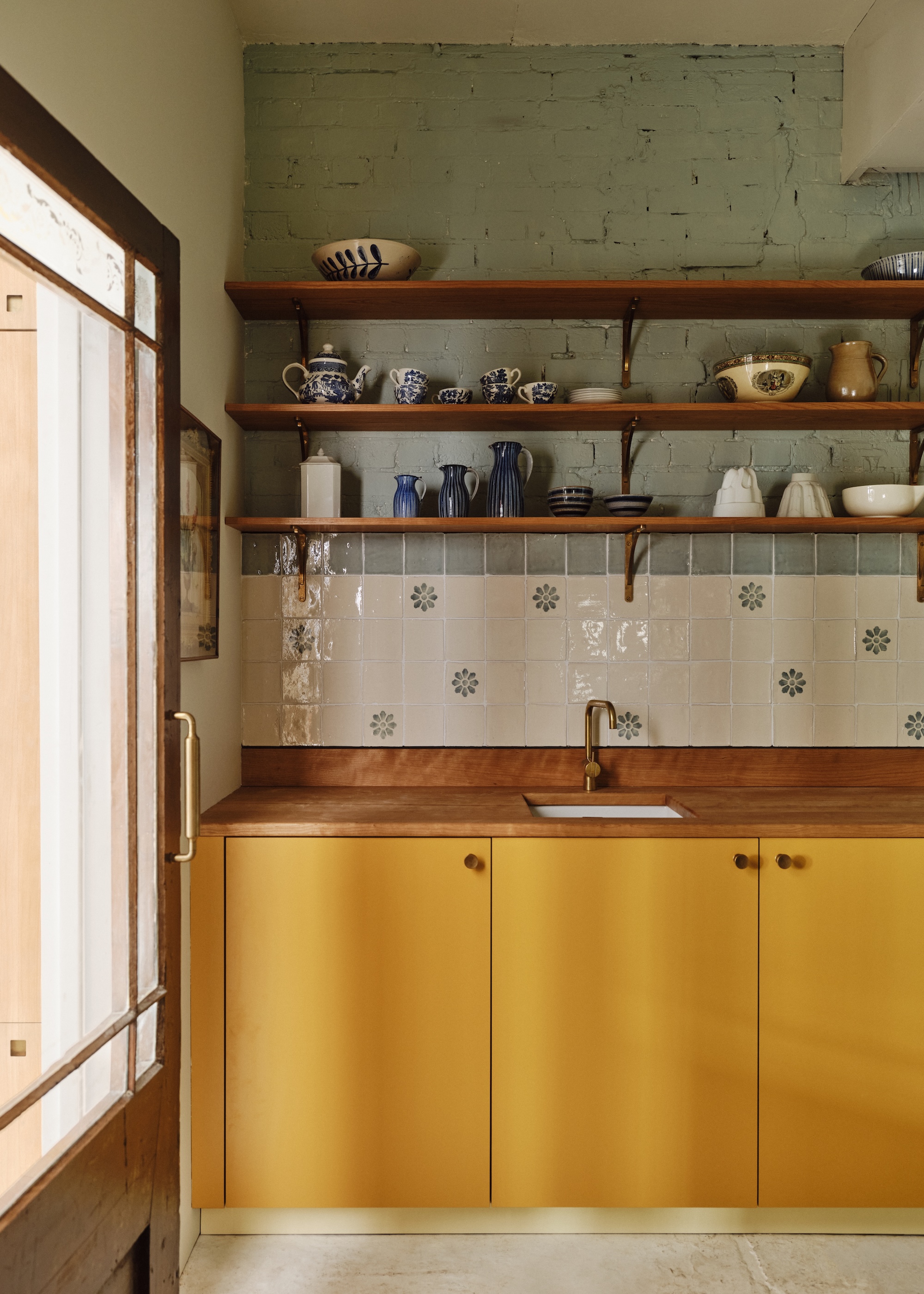a modern classic kitchen with ochre yellow base units and a backsplash with a mix of cream and sage green tiles plus oak open shelving