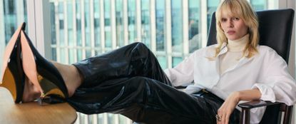 Model sat on an office chair with legs up on the table wearing a white shirt, cream turtle neck and leather trousers sold at Karen Millen