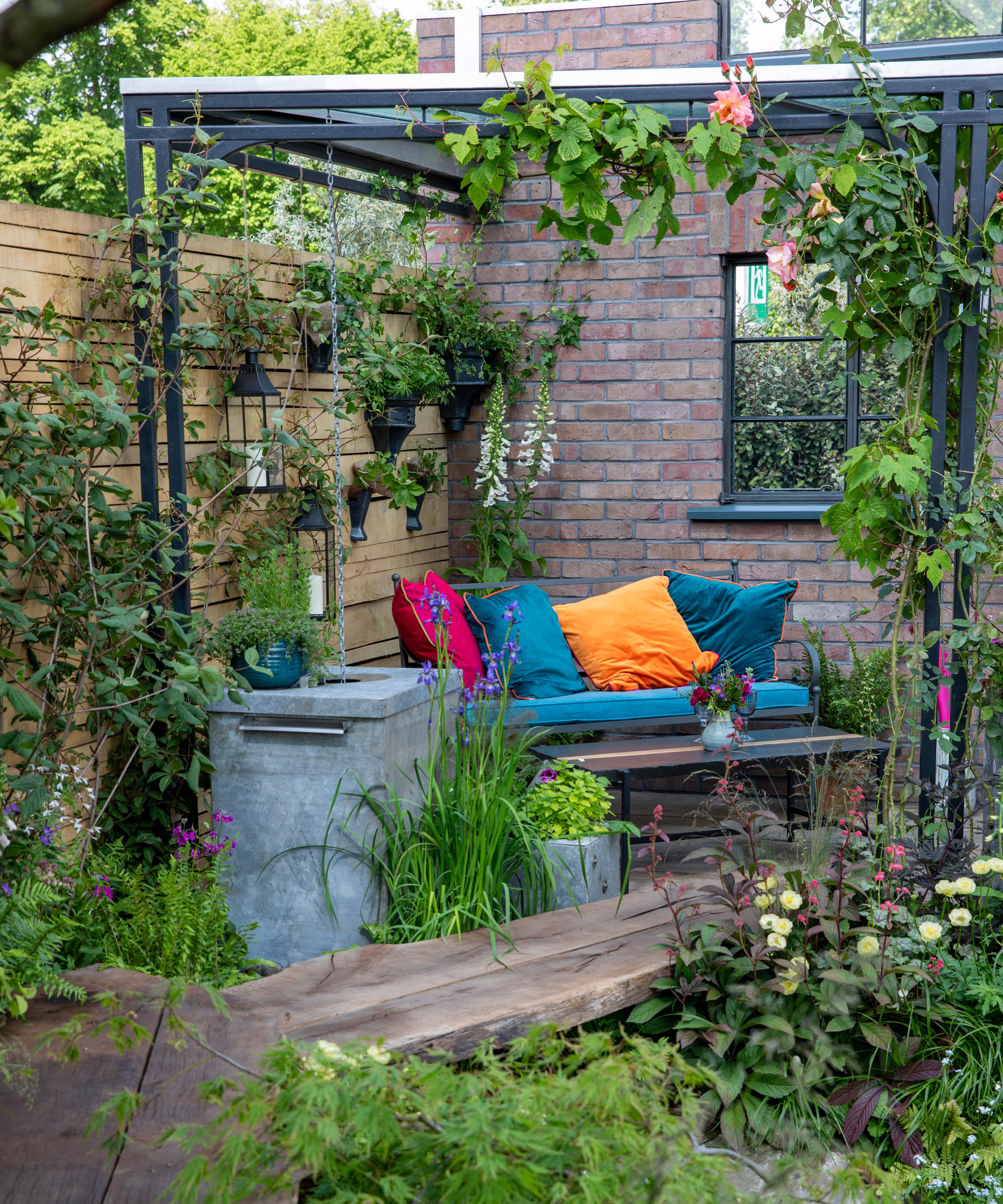front yard patio area with sofa and cushions, surrounded by a fence with climbing plants and lanterns, accessed by a path