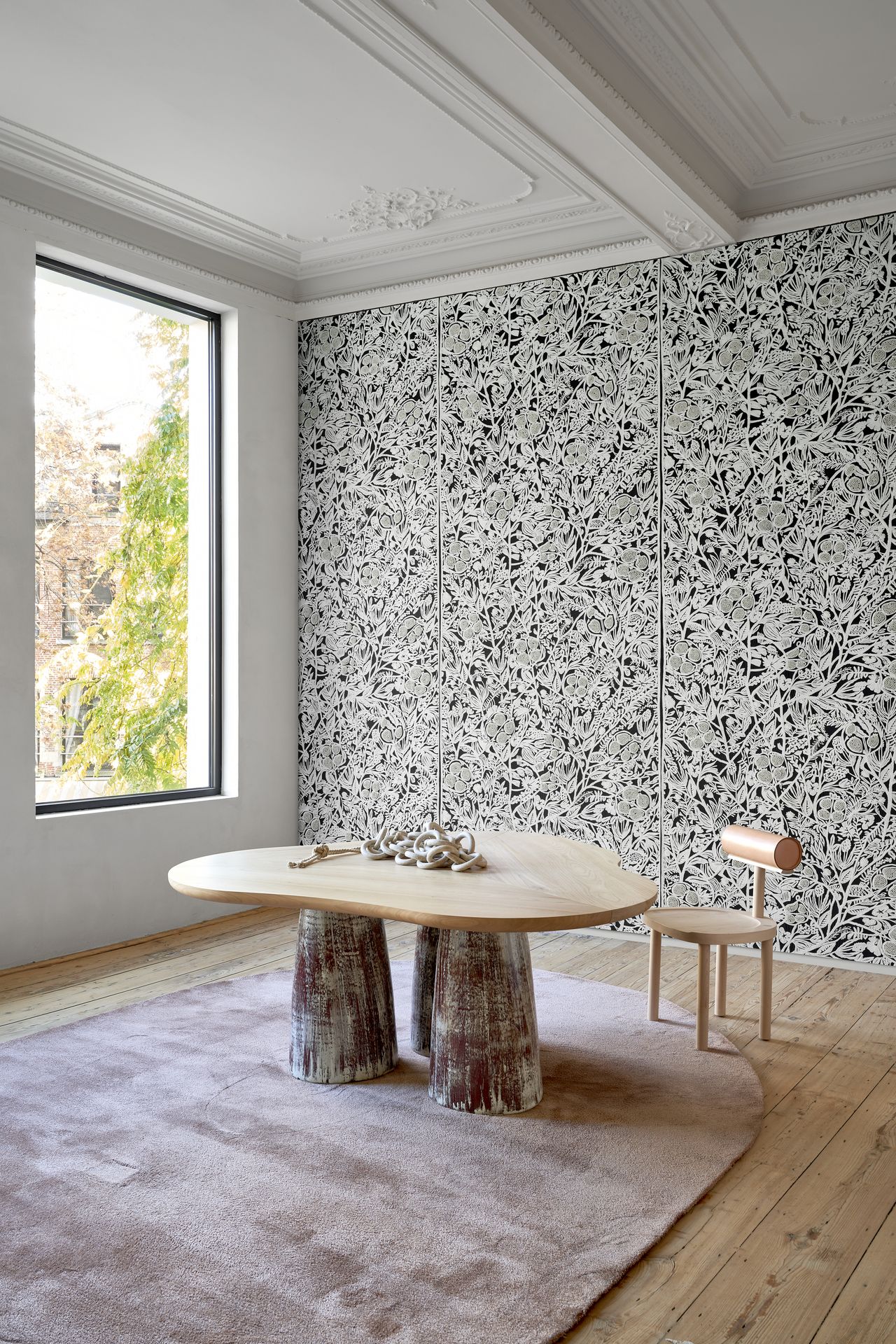 An office room with a dusty pink rug and an organically shaped wooden table. The back wall is covered in a black and white wall covering that is adorned with pearl beading.