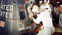 technicians in white clean room suits help an astronaut in a silver spacesuit and white helmet climb into a spacecraft for launch