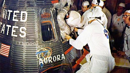 technicians in white clean room suits help an astronaut in a silver spacesuit and white helmet climb into a spacecraft for launch